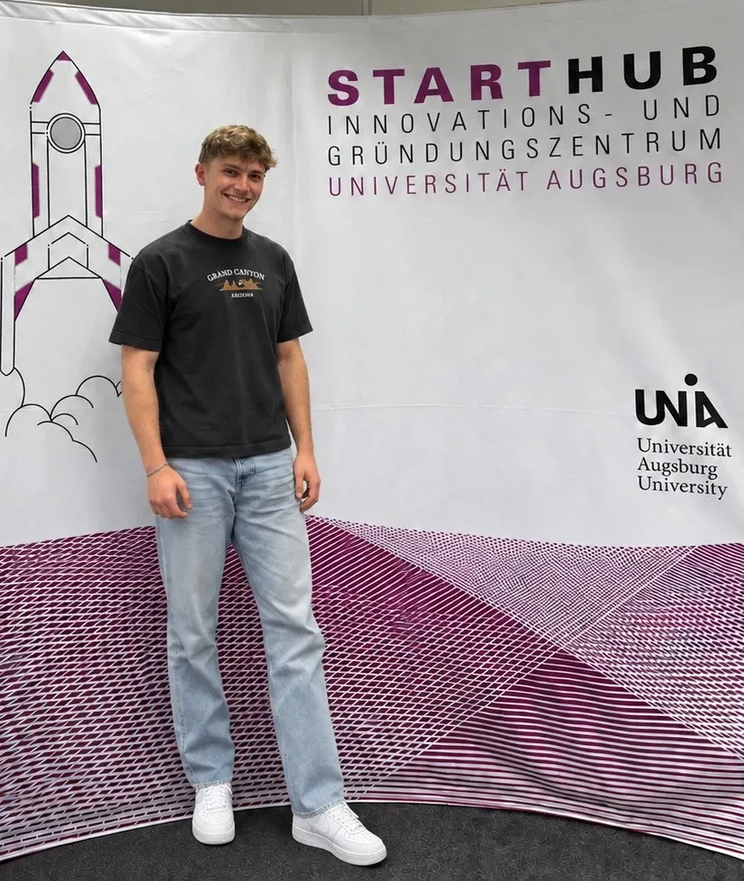 Tobias Gay vor dem Banner des StartHub, Innovations- und Gründungszentrum der Universität Augsburg (Foto mit KI bearbeitet)